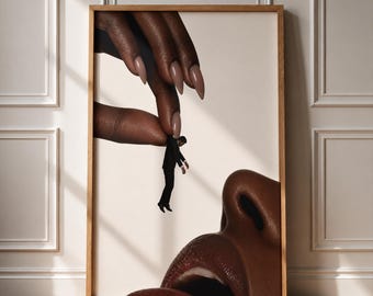 May include: A framed wall art print featuring a surreal composition of a large hand with manicured nails holding a tiny person in a black suit above a large, dark-skinned face with visible nose and lips against a cream background.
