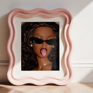 May include: A framed portrait of a doll with dark skin, long wavy brown hair, and dark lipstick, wearing black cat-eye sunglasses and small square stud earrings. The doll has its tongue sticking out. The frame is light pink with a wavy border.