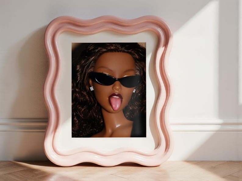 May include: A framed portrait of a doll with dark skin, long wavy brown hair, and dark lipstick, wearing black cat-eye sunglasses and small square stud earrings. The doll has its tongue sticking out. The frame is light pink with a wavy border.