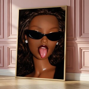 May include: A framed wall art print featuring a close-up portrait of a dark-skinned doll with wavy brown hair, wearing black cat-eye sunglasses and small square stud earrings. The doll has a glossy complexion and is sticking out its tongue.