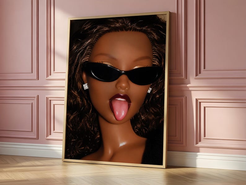 May include: A framed wall art print featuring a close-up portrait of a dark-skinned doll with wavy brown hair, wearing black cat-eye sunglasses and small square stud earrings. The doll has a glossy complexion and is sticking out its tongue.