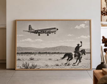 May include: A rectangular framed wall art print in black and white featuring a vintage propeller airplane flying low over a desert landscape where a person on horseback is riding and waving a hat toward the aircraft in the sky.