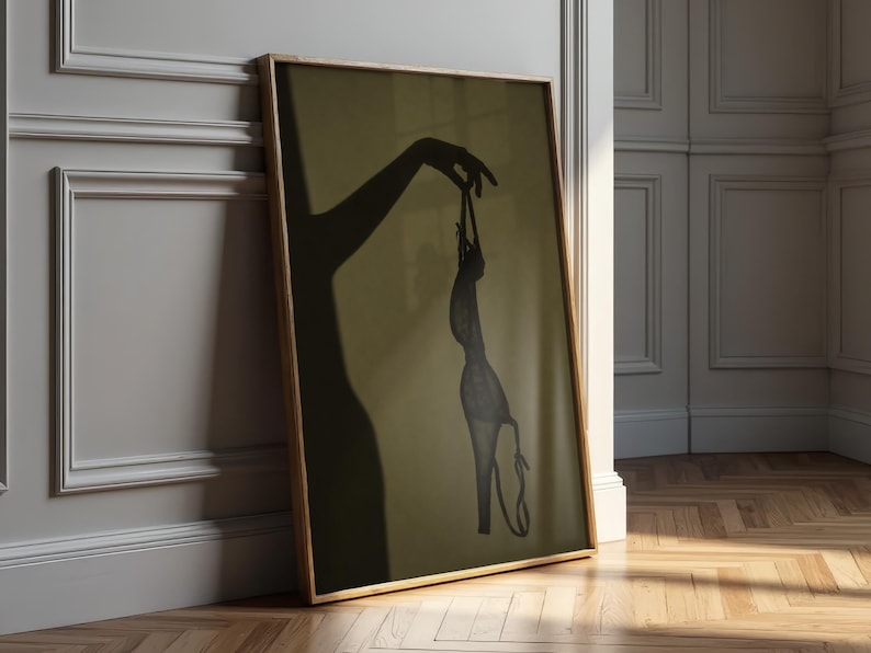 May include: A framed vertical wall art print featuring a dark silhouette of a hand holding a pair of high-heeled shoes by the straps. The artwork is set against a muted olive green background, capturing the shadow of an arm and the footwear.