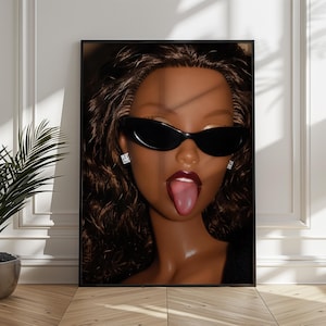May include: A framed wall art print featuring a close-up portrait of a doll with dark brown curly hair, wearing black oval sunglasses and square stud earrings. The doll has dark skin, deep red lipstick, and is sticking its tongue out.