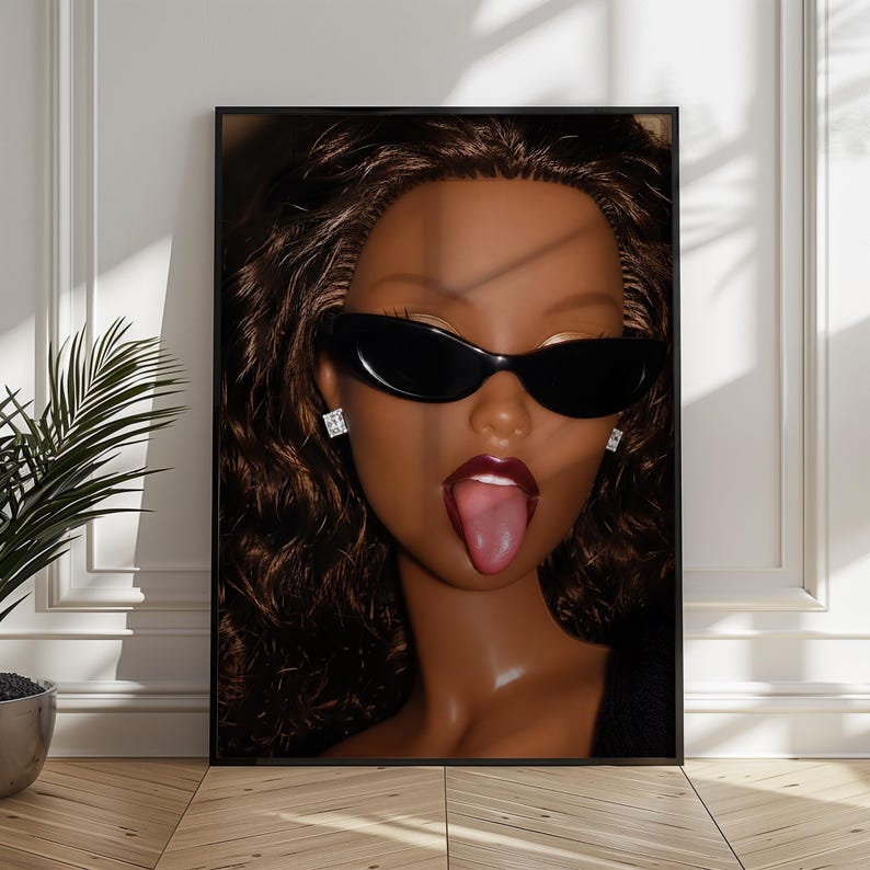 May include: A framed wall art print featuring a close-up portrait of a doll with dark brown curly hair, wearing black oval sunglasses and square stud earrings. The doll has dark skin, deep red lipstick, and is sticking its tongue out.