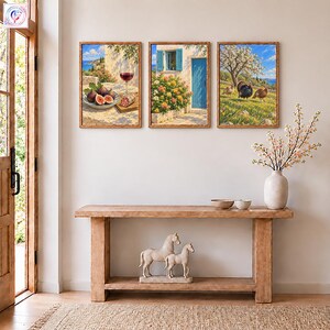 Può includere: Tre stampe da parete incorniciate raffiguranti scene mediterranee: un piatto di fichi e vino, una porta blu con fiori e tacchini in un campo. Sotto, una console in legno sostiene un vaso bianco con rami e una statuetta di cavallo.