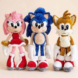 May include: Three handmade crochet plush dolls featuring characters with large eyes, pointed ears, and textured yarn bodies. The left doll is pink and red, the center doll is blue and tan, and the right doll is golden brown and white with two tails.