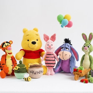 May include: A collection of crochet amigurumi figures including Tigger, Winnie the Pooh, Piglet, Eeyore, and Rabbit. Accessories include a honey pot labelled HUNN, a small bee, a wheelbarrow with vegetables, and a cluster of colourful balloons.