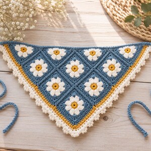 Könnte beinhalten: Ein dreieckiges Häkel-Bandana mit einem Muster aus Gänseblümchenmotiven mit weißen Blütenblättern und gelben Mitten auf blauem Grund. Die Kanten sind mit einer senfgelben Borte und einem weißen Wellenrand versehen, mit langen blauen Bindebändern.