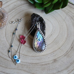 May include: A silver chain necklace with a teardrop-shaped pendant featuring a multi-colored iridescent labradorite stone. The necklace also includes two blue evil eye charms and a pink crystal charm.