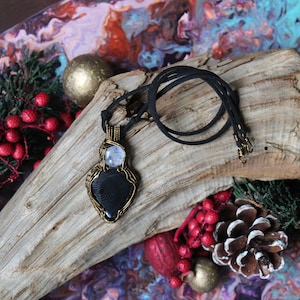 May include: A black and white stone pendant wrapped in gold wire with a clear crystal on top. The pendant is hanging from a black cord on a piece of driftwood.