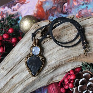 May include: A black and white gemstone pendant with a wire-wrapped design, on a black cord necklace. The pendant is on a piece of driftwood with red berries and pine cones.