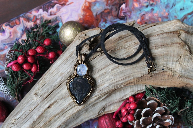 May include: A black and white gemstone pendant with a wire-wrapped design, on a black cord necklace. The pendant is on a piece of driftwood with red berries and pine cones.