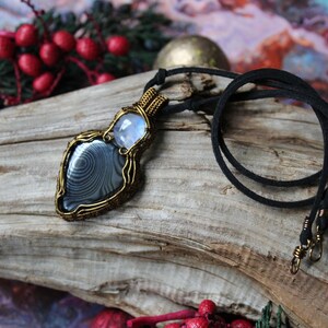 May include: A silver and black gemstone pendant with a wire-wrapped design, hanging from a black cord necklace. The pendant is on a piece of driftwood.