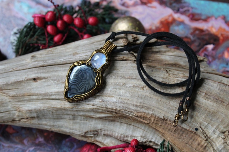 May include: A silver and black gemstone pendant with a wire-wrapped design, hanging from a black cord necklace. The pendant is on a piece of driftwood.