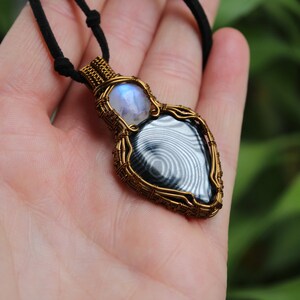 May include: A gold wire wrapped pendant featuring a black and white banded agate gemstone and a moonstone. The pendant is hanging from a black cord.