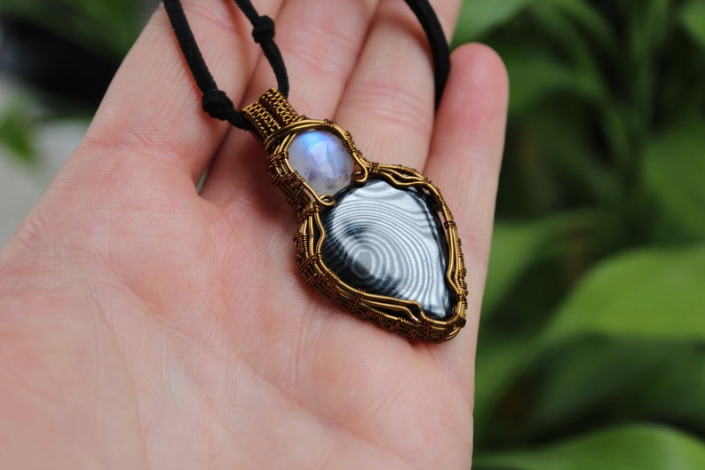 May include: A gold wire wrapped pendant featuring a black and white banded agate gemstone and a moonstone. The pendant is hanging from a black cord.