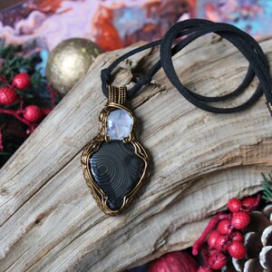 May include: A black and grey agate stone pendant wrapped in gold wire with a clear quartz stone above it. The pendant is on a black suede cord necklace.