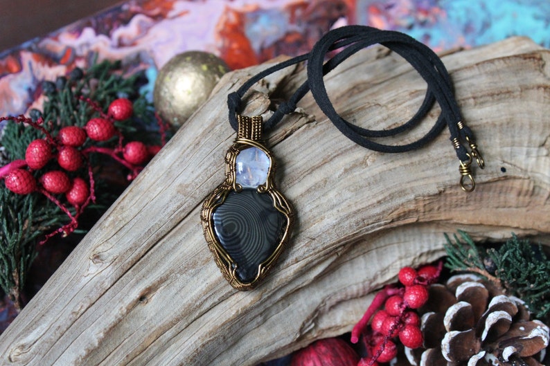 May include: A black and grey agate stone pendant wrapped in gold wire with a clear quartz stone above it. The pendant is on a black suede cord necklace.