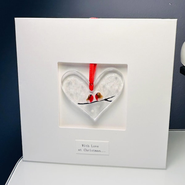 Puede incluir: Un marco cuadrado blanco que contiene un adorno de cristal en forma de corazón con una cinta roja. El adorno muestra dos pájaros pequeños con cabezas rojas posados en una rama negra sobre fondo blanco. Una etiqueta debajo dice With Love at Christmas...