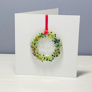 May include: A white greeting card featuring a fused glass wreath ornament. The wreath is circular, composed of green, yellow, and red glass pieces. A red ribbon attaches the ornament to the card. The card is on a white surface.