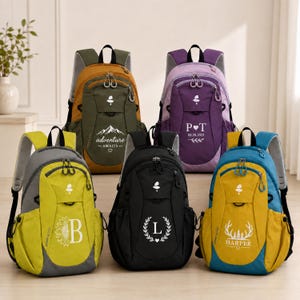 May include: Five backpacks in olive, purple, lime green, black, and mustard yellow. Each bag features a top handle, shoulder straps, and front zippered pockets. Custom white graphics include text like 'adventure AWAITS', 'P heart T', 'B', 'L', and 'HARPER'.