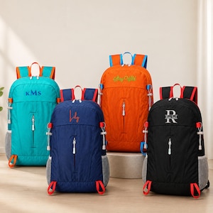 Puede incluir: Cuatro mochilas en turquesa, azul marino, naranja y negro, cada una con correas ajustables, bolsillos laterales de malla, un compartimento frontal vertical con cremallera y bordados personalizados con los nombres KMS, Lily, Rachel y el texto Stay Wild.