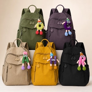 May include: Five backpacks in olive green, dark purple, beige, mustard yellow, and black. Each bag features a front zippered pocket, side pockets, and a unique crocheted letter or shape charm with googly eyes and dangling limbs attached to the zipper.