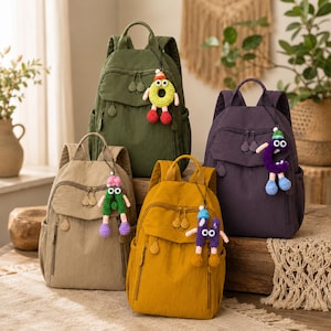 Puede incluir: Cuatro mochilas en verde oliva, morado oscuro, beige y amarillo mostaza, cada una con asa superior, bolsillo frontal con cremallera y un dije de personaje tejido a ganchillo con ojos móviles y extremidades colgantes en la cremallera.