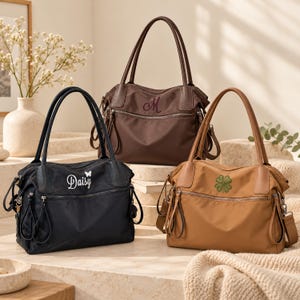 May include: Three nylon tote bags in black, brown, and tan. The black bag features the text Daisy with a small butterfly. The brown bag has a script letter M, and the tan bag displays a green four-leaf clover. Each bag has dual handles.