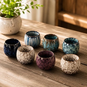 Può includere: Un set di sette piccole tazze da tè in ceramica di forma rotonda con varie finiture in smalto reattivo. La collezione include tazze blu scuro, bianche con macchie marroni, azzurre con striature verticali, turchesi, viola e verde acqua con texture.