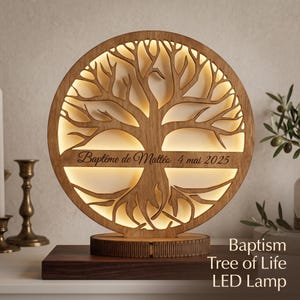 Può includere: Una lampada LED circolare in legno con un design ad albero della vita tagliato al laser. Una barra orizzontale in legno al centro mostra il testo inciso Baptême de Mattéo 4 mai 2025. La lampada emette una luce calda dietro la struttura.