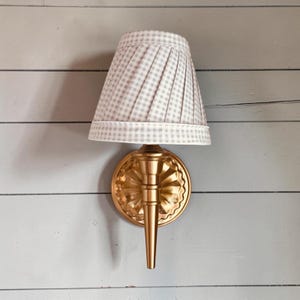 Könnte beinhalten: Eine Wandleuchte mit goldfarbenem Metallfinish, einer runden, gewellten Wandplatte und einem konischen Arm. Die Leuchte ist mit einem plissierten, kegelförmigen Stoffschirm ausgestattet, der ein hellgrau-weißes Vichy-Karo-Muster aufweist.