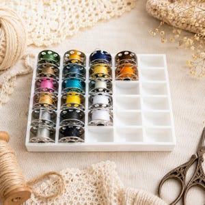 Peut inclure: Un plateau de rangement en plastique blanc contenant plusieurs canettes de machine à coudre en métal enroulées de fils de diverses couleurs, dont vert, jaune, bleu, rose, orange, blanc, noir et argent, disposées en rangées dans les compartiments.