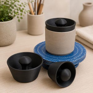 May include: A set of three matte black and beige containers with lids. One container is beige with a black lid, while the other two are black bowls with central knob handles. The items are arranged together, showcasing a cylindrical and bowl-like design.
