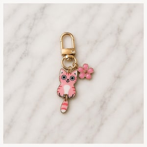 Puede incluir: Un llavero de metal dorado con un dije de esmalte rosa y blanco en forma de gato con rayas y grandes ojos negros, acompa&ntilde;ado de un peque&ntilde;o dije de flor rosa de cinco p&eacute;talos unido a una argolla circular y un cierre de mosquet&oacute;n.