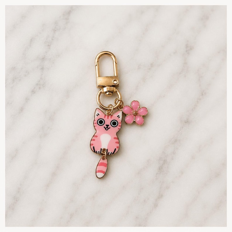 Puede incluir: Un llavero de metal dorado con un dije de esmalte rosa y blanco en forma de gato con rayas y grandes ojos negros, acompa&ntilde;ado de un peque&ntilde;o dije de flor rosa de cinco p&eacute;talos unido a una argolla circular y un cierre de mosquet&oacute;n.