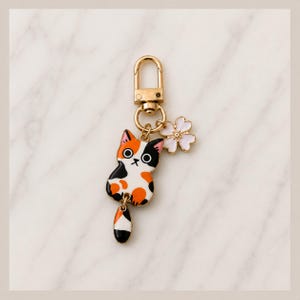 Puede incluir: Un llavero de metal dorado con un dije de gato calic&oacute; con parches de esmalte naranja, negro y blanco. El gato tiene una pieza de cola colgante separada y se acompa&ntilde;a de un peque&ntilde;o dije de flor blanca de cinco p&eacute;talos con centro dorado.
