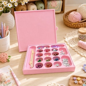 May include: A square pink plastic storage organizer with a hinged lid, featuring multiple circular and oval compartments filled with assorted colorful beads and small wooden tools, designed for organizing craft supplies and jewelry components.