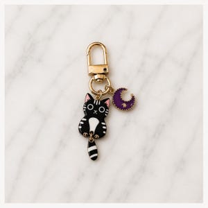 Puede incluir: Un llavero de metal dorado con un dije de esmalte negro y blanco que representa un gato con el pecho blanco y un dije separado de media luna morada con una peque&ntilde;a estrella dorada, unido a un cierre de mosquet&oacute;n y una argolla circular.