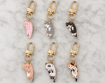 Puede incluir: Seis llaveros de metal con dijes de gato de esmalte en varios colores, incluyendo rosa, gris, blanco y negro, naranja, siamés y calicó. Cada dije cuelga de un cierre de mosquetón de tono dorado y una pequeña argolla circular.