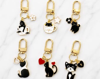 Puede incluir: Seis llaveros de metal dorado con diseños de gatos en blanco y negro. Cada llavero incluye un cierre de mosquetón y varios dijes, incluyendo un gato en una taza, un gato con un corazón, una estrella y un candado, con detalles en esmalte.