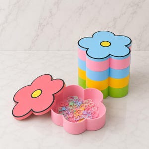 May include: A stackable flower-shaped storage container with four colorful tiers in blue, pink, yellow, and green. The top lid is pink with a yellow center. A separate pink flower-shaped tray contains a collection of small, pastel-colored plastic rings.