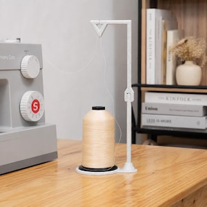May include: A white speckled 3D-printed thread stand with a vertical arm and a triangular top guide, holding a large spool of beige sewing thread. The stand is positioned next to a grey sewing machine with two white adjustment dials on the front.