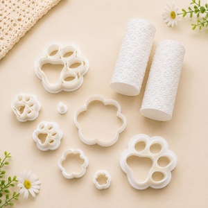 Può includere: Un set di formine in plastica bianca per argilla e due mattarelli cilindrici. Il set include formine a forma di impronta di zampa di varie dimensioni, una formina a fiore e piccoli tagliapasta circolari con texture.