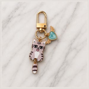 Puede incluir: Un llavero de metal dorado que presenta un dije de gato a rayas de color morado claro y blanco con una cola colgante, junto con un peque&ntilde;o dije de coraz&oacute;n turquesa adornado con alas doradas, unido a un cierre de mosquet&oacute;n y una argolla.