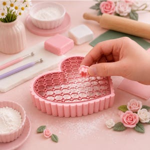 May include: A heart-shaped pink plastic mold with a honeycomb grid pattern, used for crafting small shapes. A hand holds a tiny matching pink flower-shaped cutter above the mold. The scene includes small bowls of white powder, sculpting tools, and roses.