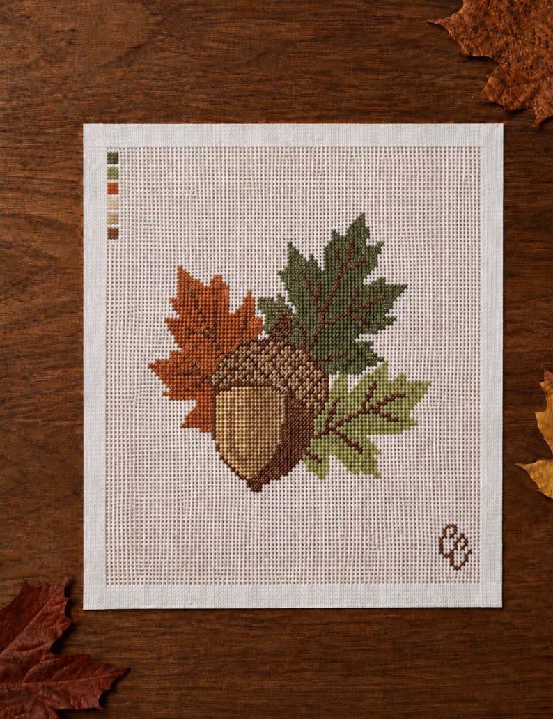 May include: A cross-stitch pattern on white fabric featuring a central brown acorn surrounded by three maple leaves in shades of orange, dark green, and light green. A small color palette guide is in the top left corner and a logo is in the bottom right.