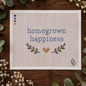 May include: A white needlepoint canvas featuring the blue text homegrown happiness, centered above two green leafy branches and a tan heart. A small color palette of blue, green, and tan squares is in the top left corner, with a logo in the bottom right.