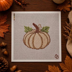 Peut inclure: Une toile de canevas carrée présentant un motif au point de croix représentant une citrouille beige clair avec une tige brune et deux feuilles vertes. La toile a un fond quadrillé blanc, un petit guide de couleurs en haut à gauche et des initiales en bas à droite.